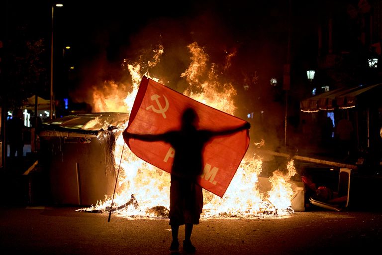 Протестующий держит флаг рядом с горящими мусорными баками во время столкновений с полицией Протестующий держит флаг рядом с горящими мусорными баками во время столкновений с полицией