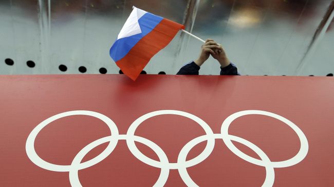 Болельщик держит российский флаг над логотипом олимпийских игр. Архивное фото Болельщик держит российский флаг над логотипом олимпийских игр. Архивное фото